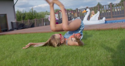 Young girl doing somersaults on the grass in the backyard of a country or suburban house. The child is having fun by the pool on a hot sunny day. 4k uhd slow motion footage