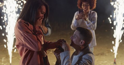Cinematic shot of homosexual female gay is making surprise proposal of marriage to beloved woman on background of happy friends dancing with sparklers during engagement celebration party at night.
