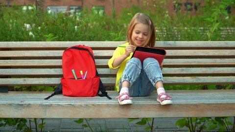 Little child sits on bench with backpack in schoolyard and does homework. Student girl draws outdoors. Education and school concept.
