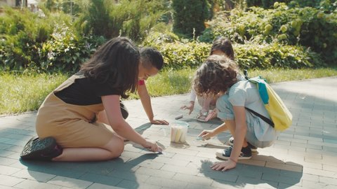 Group of lovely mixed race school children making sidewalk chalk painting at sunny day in public park. Diverse elementary students involved in fun summer activity, drawing with crayons on pavement