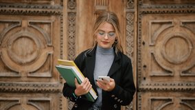 Student girl in glasses with books walking to the university building. Portrait of attractive girl texting on her phone.