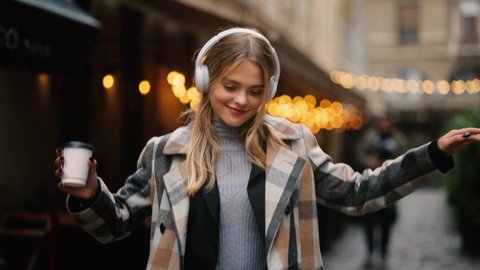 Slow motion of smiling young blond listening to music in headphones at the city street. Closeup face of beautiful young girl with long hair.
