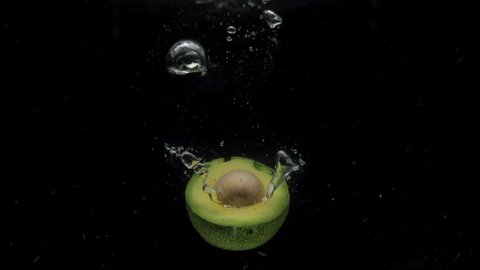 Slow motion one avocado halves with seed falling into transparent water on black background. Fresh fruits splashing in aquarium. Healthy food, diet, air bubbles