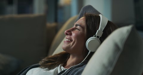 Cinematic shot of young happy smiling woman is lying on sofa with headphones and relaxing with comfort while listening her favorite music playlist in living room at home with soft atmospheric light.