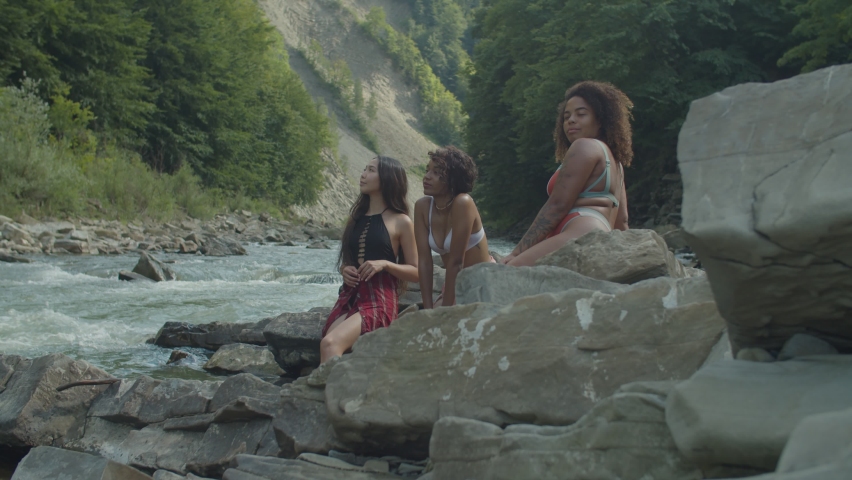 Top view of charming relaxed multiracial females in swimsuits sitting on rocks, dangling feet into water, sunbathing, enjoying scenic nature, recreation and freedom while resting at mountain river.