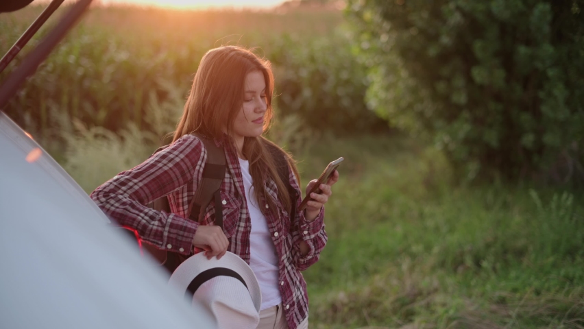 Young attractive woman traveler uses smartphone while standing near car at sunset. Steadicam shot.