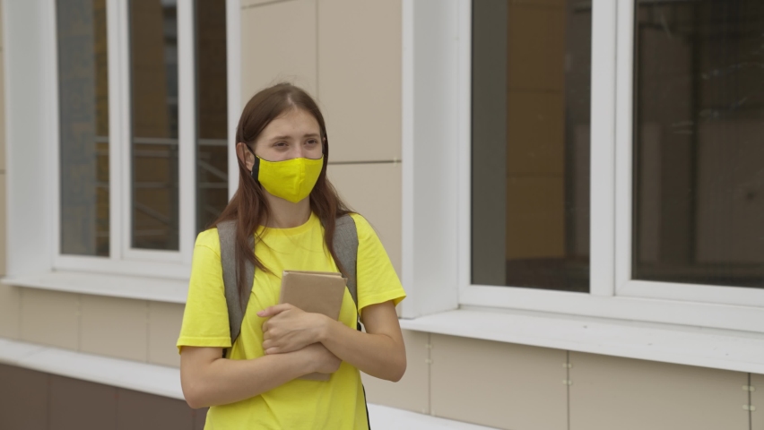 A happy student in a protective mask with a book in her hand goes to college, teenager in the schoolyard, cheerful girl loves to read, go to library during pandemic, new different life of coronavirus Royalty-Free Stock Footage #1079469023