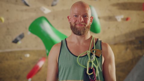 Strong Masculine Male Athlete Smiling and Posing at Rock Climbing Gym with Bouldering Wall Background. Handsome Happy Bald Man with Ginger Beard, Wearing Colorful Undershirt and Holding Rope Harness.