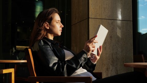 Beautiful girl sits with open book in urban street cafe. Young woman spends time reading while waiting for somebody in city. Lady booklover, student, philologist, reader, bookworm