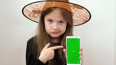 Little girl in witch Halloween costume shows the phone with a green screen. Chroma key green screen smartphone set up for advertising. Cute child looking at camera. Adorable kid with digital device 