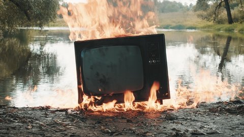 Retro vintage TV flares up and burns with fire next to the river. Concept of post apocalypse, surrealism and the fire hazard of old worn out equipment