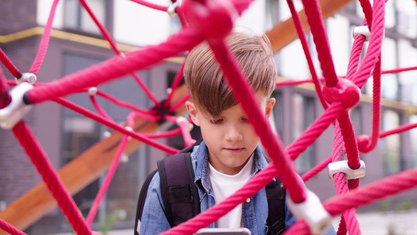 Friendly child with backpack sitting on the school playground after lessons. Waiting for his parents to come after him, playing games on phone. Back to school concept. Royalty-Free Stock Footage #1079486627