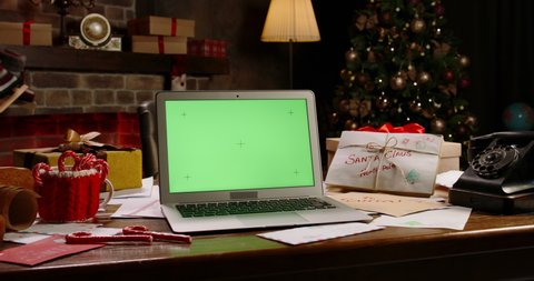 Laptop with green screen at Santa Claus workplace. Close-up of computer with chroma key on table. Christmas time, holidays and celebrations concept. New Year tree and fireplace on background.