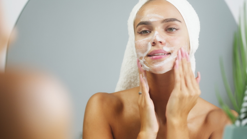 Woman using moisturizing foam for face washing and smiling, morning skincare routine. Young female student wearing towel on head looking in mirror in spa salon. Preparation for make up. | Shutterstock HD Video #1079489417