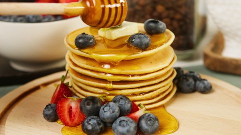 Pouring honey or maple syrup on pancakes with berries on top close-up. Fresh cooked blueberries and strawberries flapjacks for breakfast. Stack of oatmeal pancakes. Healthy vegetarian sweet food. 