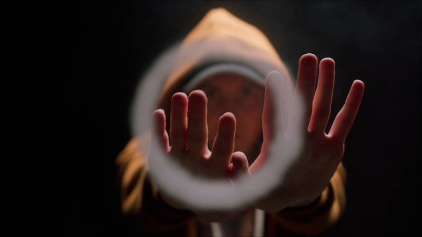 Close-up of young man wearing hoodie and hat blowing out smoke rings, guy smoking hookah or cigarette in dark studio, puffs of smoke from the mouth on black background. Bad habits concept.  | Shutterstock HD Video #1079489606