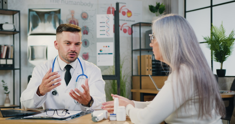 Focused physician in medical coat sitting at table consulting middle aged female patient about illness or surgery Royalty-Free Stock Footage #1079491322