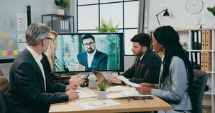 Attractive positive experienced businesspeople have video conference with their male colleague on tv during meeting in modern boardroom