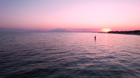 Pull back view of pink, summer sunset. Young man fishing in the backrgound. Pink reflections of the sky in the wavy waters. : vidéo de stock