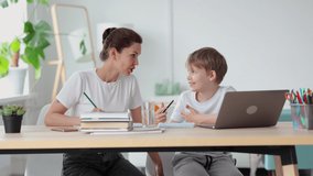 Mom, son talking and using laptop while learning online at table in home room. Front view of young woman, boy talk with smiles and look at computer screen, sit at desk in interior. American