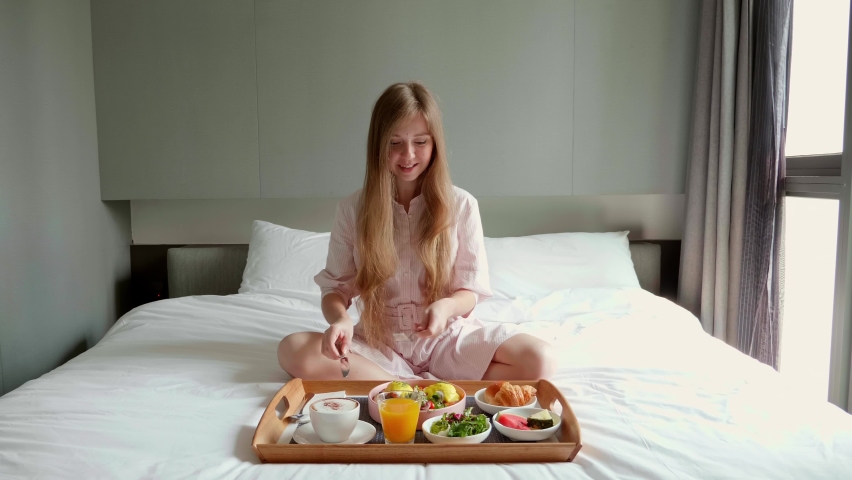 Smiling Woman Sitting on Bed with Wooden Tray Full of Food, Eating Fresh Food. Breakfast in Bed in Hotel. Healthy Morning Eating with Fresh Eggs, Fruits and Vegetables on Vacation. Room Service. 4k