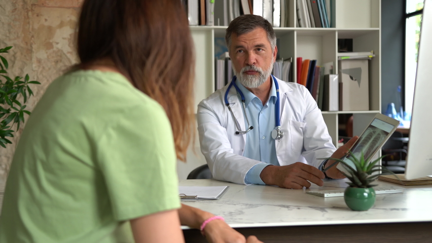 Serious focused aged physician in medical coat sitting at table, consulting female patient about illness or surgery Royalty-Free Stock Footage #1079497562