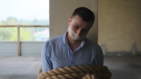 businessman tied with rope with his mouth sealed with tape sits tied to post, looks up at camera. Seen man in blue shirt, victim of maniac with gag in his mouth, cannot speak, dumb.