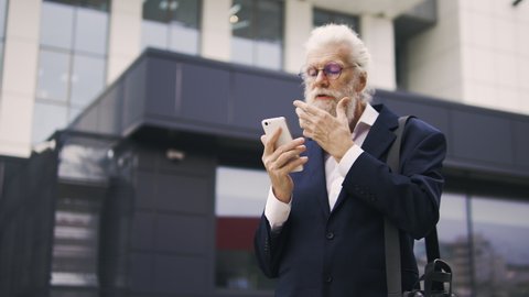 Old stylish man adjusting his long white beard looking on smartphone display