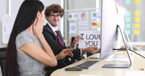Businesspeople secret love affair and flirting in office concept. Male and female colleagues sitting and working with computer, man raises paper with 'i love you' words to woman show feeling in mind.
