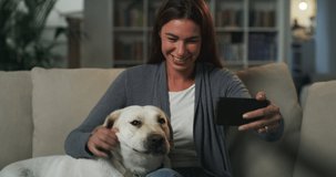 Portrait of a Woman Sitting on a Couch with her Labrador Retriever Dog, Using Smartphone and Having a Video Call. Young Female Staying Connected to Family and Friends After Moving Thanks to Internet