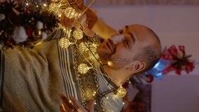 Vertical video: Festive couple knotted in string of christmas lights. Man and woman laughing while trying to untangle garland with twinkle lights and illuminated bulbs for holiday celebration.