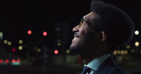 Cinematic close up shot of handsome afro american successful elegantly dressed businessman with glasses is looking around satisfied with his deal in busy city center with traffic with night lights.