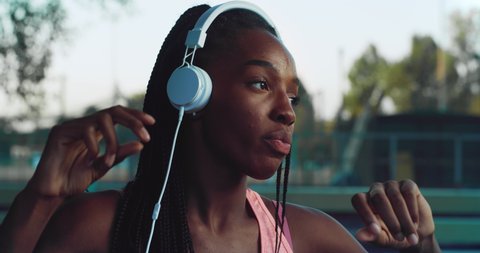 Cinematic shot of young african woman is having fun to listening to the music with earphones and dancing on basketball outdoor court after finishing friendly game match at sunset.
