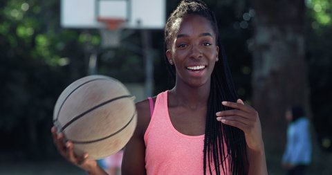 Cinematic shot of young happy african woman with dreadlocks carrying ball and smiling satisfied with her results in camera on basketball outdoor court background after finishing a friendly game match.