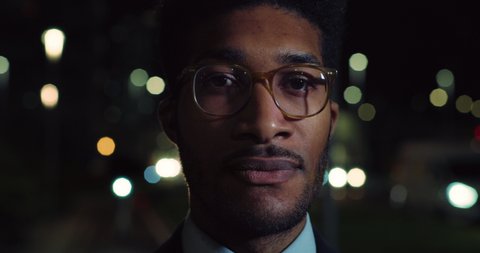 Cinematic close up shot of handsome afro american successful elegantly dressed businessman with glasses is looking around satisfied with his deal in busy city center with traffic with night lights.