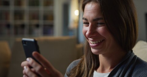 Cinematic close up shot of young smiling woman is using smartphone for entertainment while sitting on sofa in living room at home with soft atmospheric light.