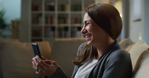 Cinematic shot of young smiling woman is using smartphone for entertainment while sitting on sofa in living room at home with soft atmospheric light.