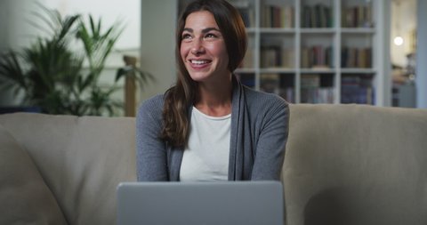 Cinematic shot of young business woman is smiling satisfied with her work while doing smart working with laptop from home on sofa in living room during lockdown with daylight.