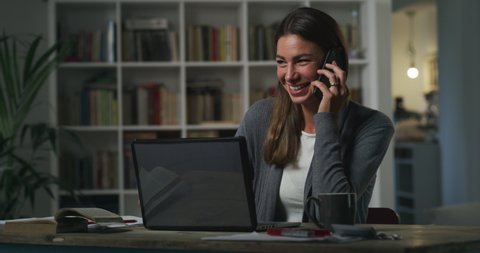 Cinematic shot of young smiling business woman is speaking by phone with friend or colleague while doing smart working with laptop from home in living room during lockdown with soft atmospheric light.