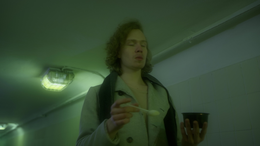 A young Caucasian man with long hair in a white coat and scarf walks through tunnel of the underpass and eats soup with a spoon from a plastic plate