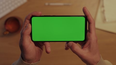 Top view of male hands holding modern smartphone in horizontal position and sliding with finger on green empty screen. Mobile application for work at home.