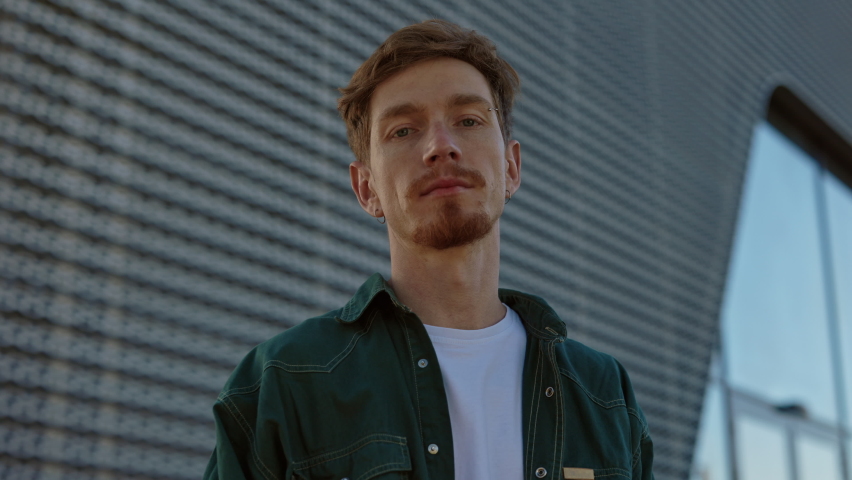 Redhead man with serious facial expression posing on camera outdoors. Bearded guy with piercing in brow and ears standing near modern building. Portrait of male hipster at urban area.