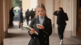 Student girl in glasses with books standing near the university building. Portrait of attractive girl texting on her phone.