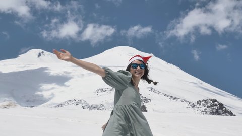 Young woman in Santa hat sunglasses and grey summer dress spins with smile dancing against snowy mountains in sunny weather