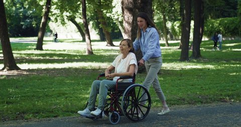 Cinematic shot of carefree happy granddaughter and grandmother with disability who uses wheelchair having fun to walk together in green park. 