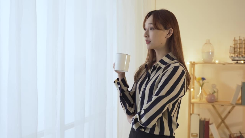 waist up asian female manager is drinking tea from cup and heaving a sigh of relief while standing by window looking outside in the home interior. Royalty-Free Stock Footage #1079542961