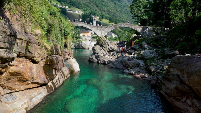 Drone shot of Ponte dei Salti the double-arched medieval stone pedestrian bridge over the clear water of the Verzasca River. In Lavertezzo in Switzerland with tourist on the bridge.