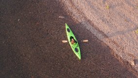 Aerial FPV fly up video from girl with red long hairs kayaking and stops at island shore to take photos of scenic Northern nature. Blue water, red sand and stones. 
