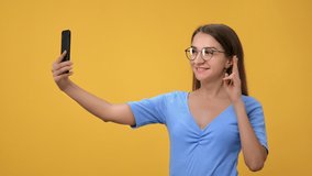 Happy young blonde woman in eyeglasses posing taking selfie use smartphone peace greeting gesture isolated on orange studio background. Smiling business lady photographing video call on mobile phone