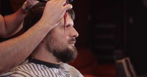Close-up of a handsome Caucasian guy sitting in a hairdressing salon. An unrecognizable barber parses hair with a comb. 